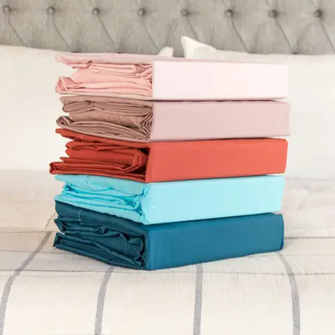 A stack of folded sheet sets in pink, mauve, rust, sky blue and teal on a bed with a grey tufted headboard