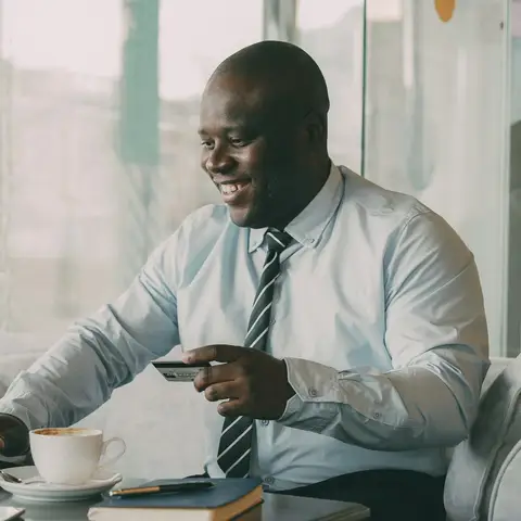 A smiling man in a shirt and tie holding a payment card while working on a laptop in a cafe