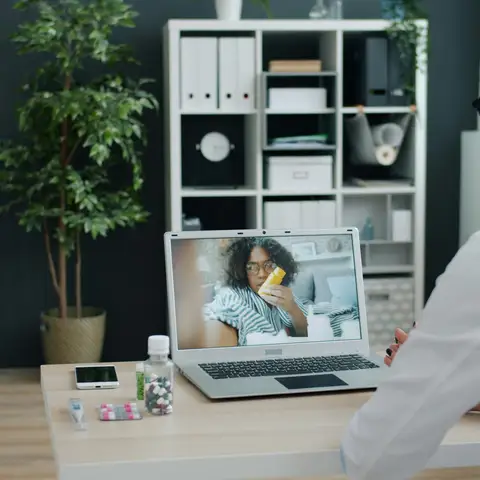 A clinician at her office desk holding a video visit on a laptop with a patient who is showing a pill bottle to the camera