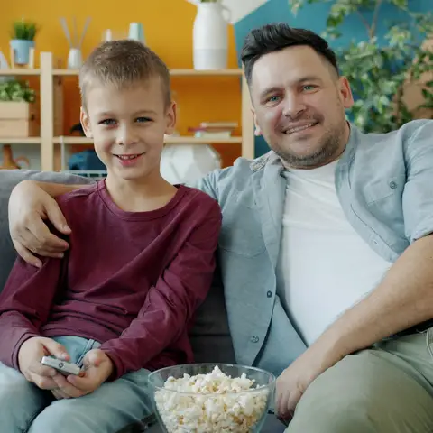 A father and his young son smiling on the sofa at home with a remote and a bowl of popcorn