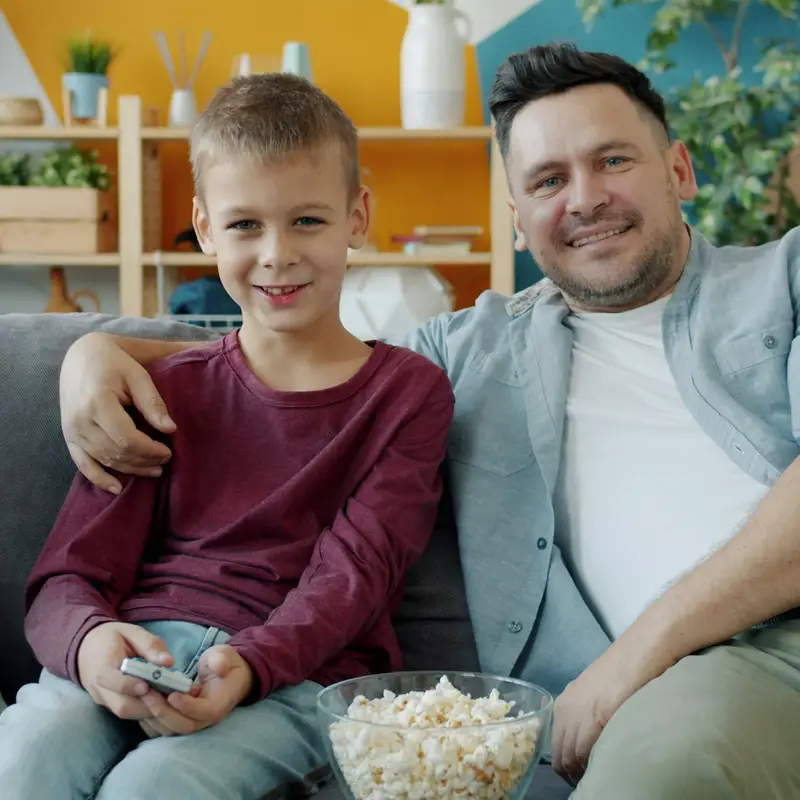Email header photo: a father and his young son smiling on the sofa at home with a remote and a bowl of popcorn
