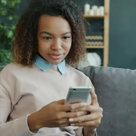 A smiling young woman sitting on a gray couch at home, scrolling on her smartphone