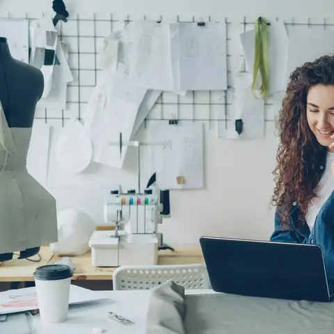 A small business owner smiling on the phone while working on a laptop in her design studio