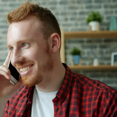 A smiling man in a plaid shirt talking on his cell phone in a room with a brick wall