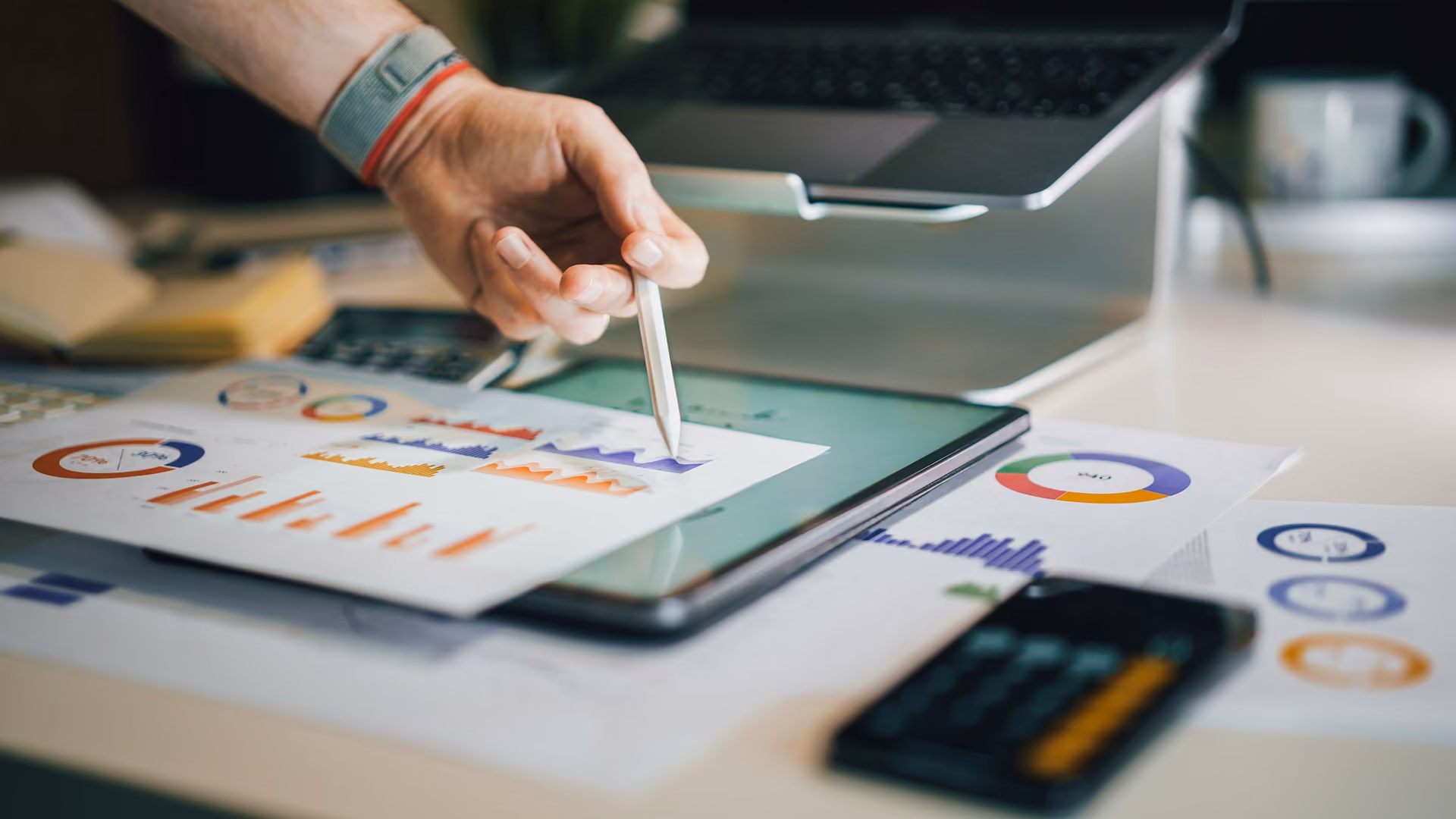 Hand with a stylus reviewing channel spend charts on a tablet, with printed reports and a calculator on the desk