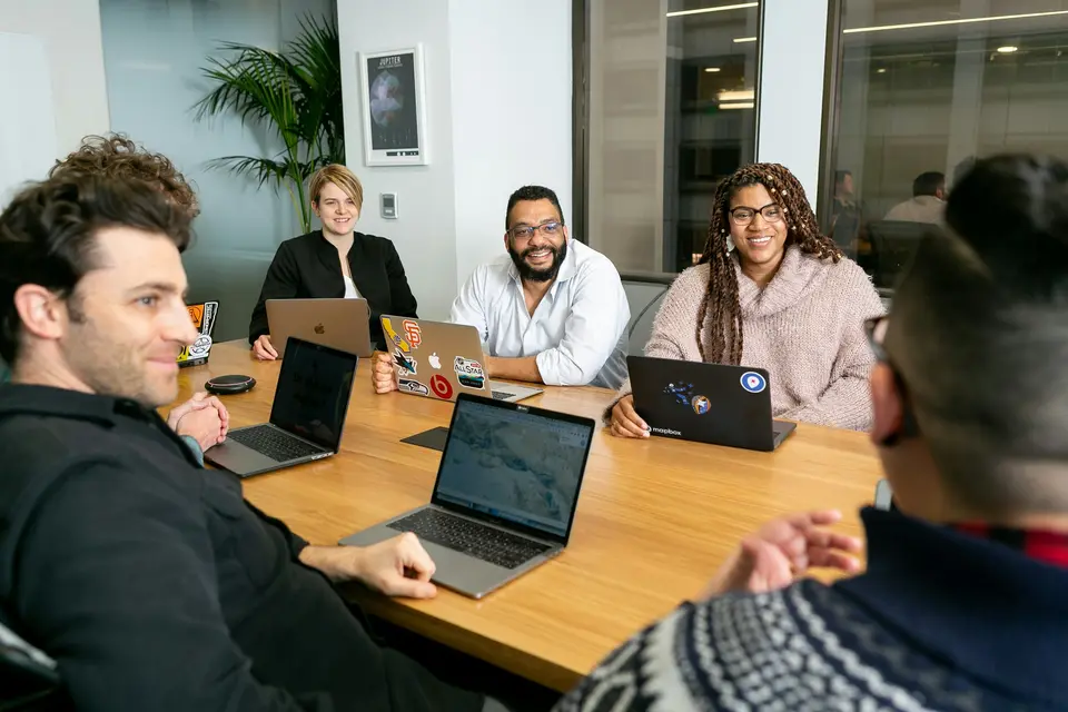 Team of four professionals listening to a colleague in a board meeting with laptops open