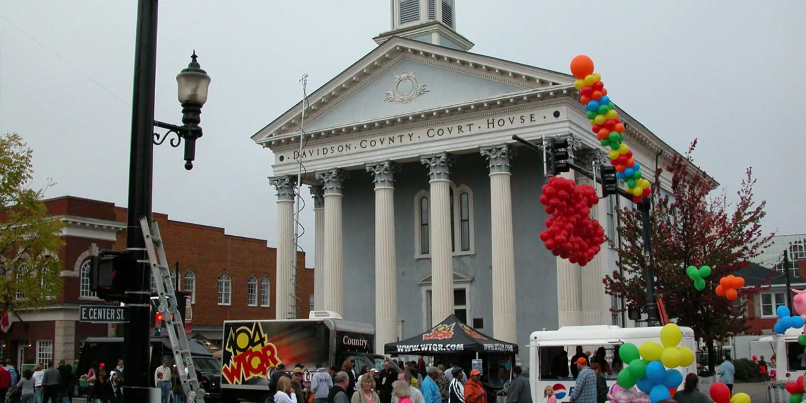Community festival crowd gathering on downtown street corners lined with historic architecture and artisan booths