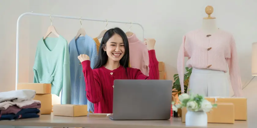 Clothing shop owner smiling at her laptop beside a rail of shirts