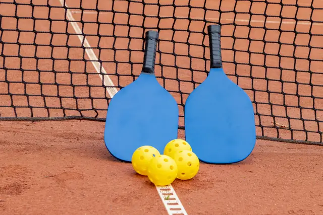 Two blue pickleball paddles and three yellow balls resting by the net on a clay court