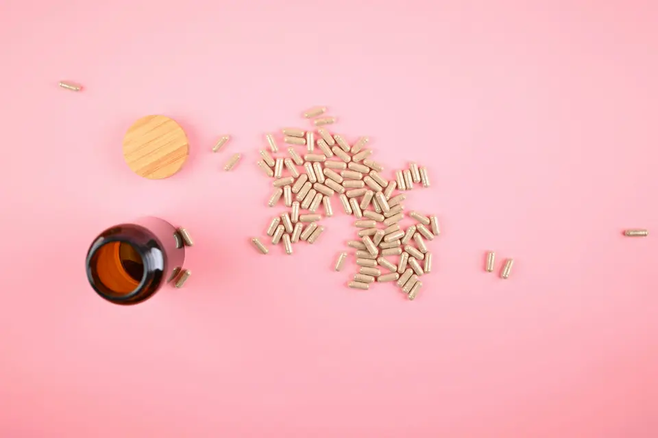Supplement capsules spilled beside a small amber bottle and a wooden lid on a pink surface