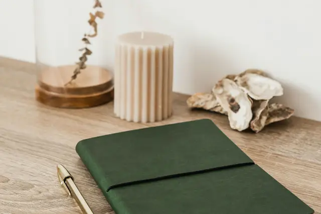 Green cloth journal and a gold pen on a light wooden desk beside a candle