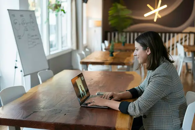 Woman in a plaid blazer on a video call at a laptop on a long wooden table