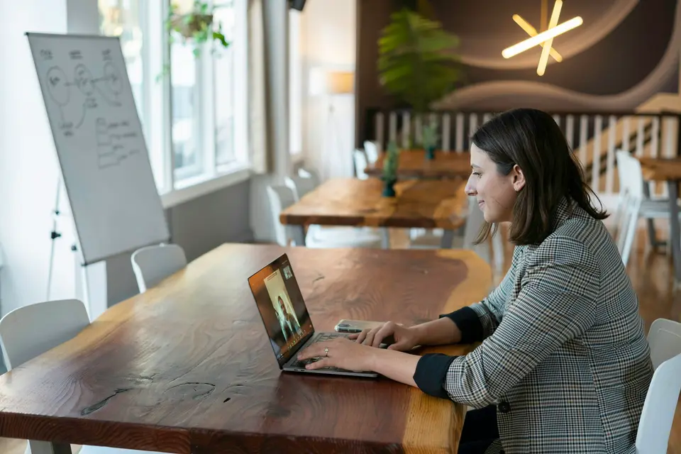 Woman in a plaid blazer on a video call at a laptop on a long wooden table