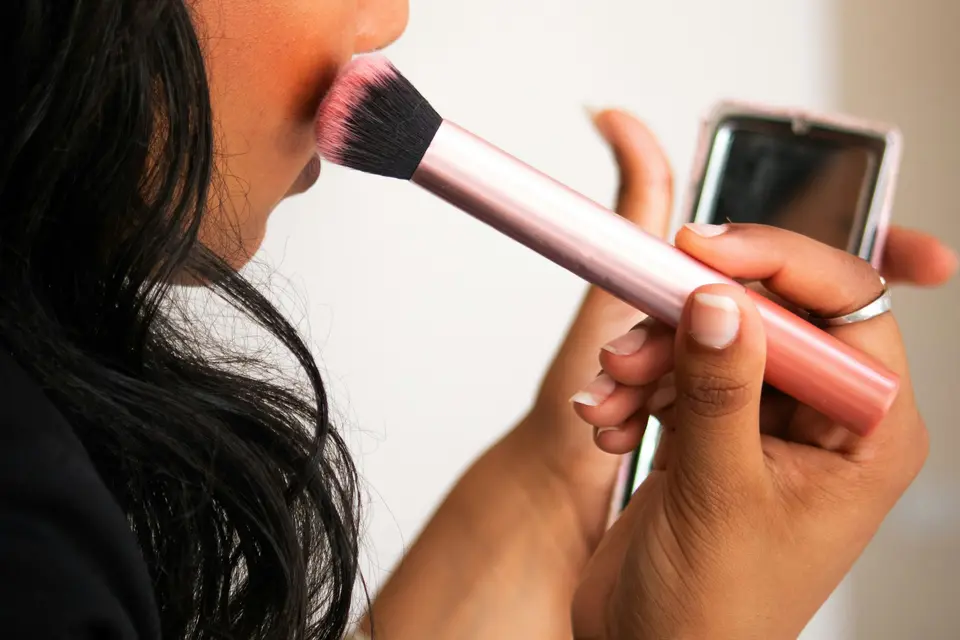 Woman applying blush to her cheek with a pink makeup brush while holding a small mirror