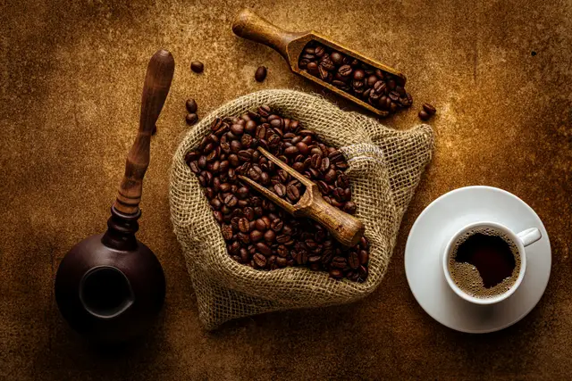 Roasted coffee beans in a burlap sack with a wooden scoop beside a cup of coffee