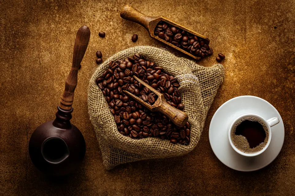 Roasted coffee beans in a burlap sack with a wooden scoop beside a cup of coffee