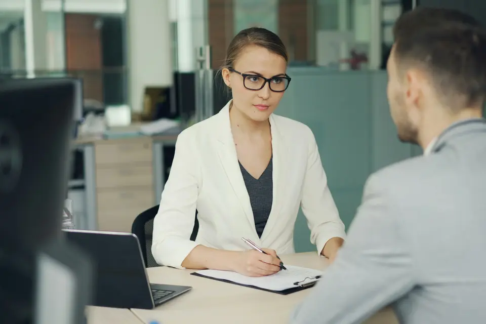 Recruiter in a white blazer taking notes while interviewing a candidate across a desk
