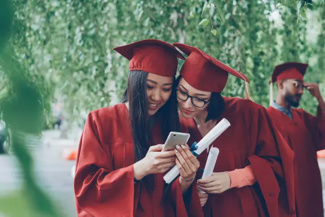Two graduates in red gowns smiling at a phone together after a ceremony
