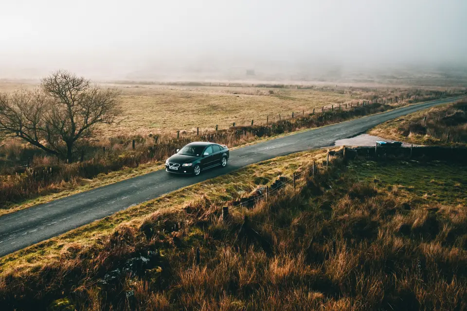 Dark sedan driving a winding country road through autumn grassland on a misty morning