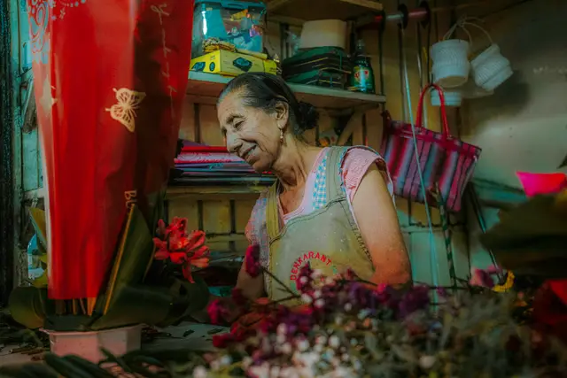 Smiling older woman in an apron arranging flowers in a small, crowded flower shop