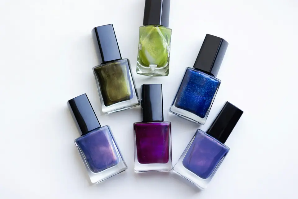 Six glass bottles of nail polish in green, blue and purple shades with black caps on a white surface