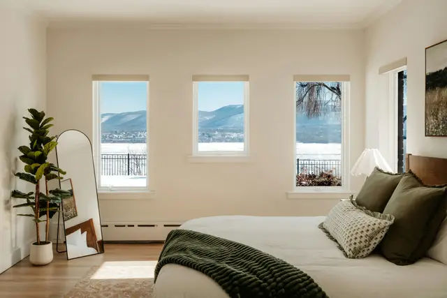Bright bedroom with a freshly made bed, green pillows and throw, and three windows looking out on snowy hills