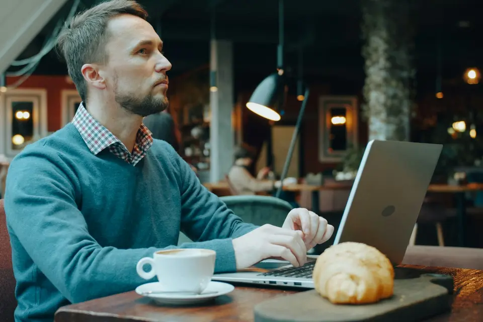 Man in a blue sweater typing on a laptop at a cafe table with a cup of coffee and a croissant