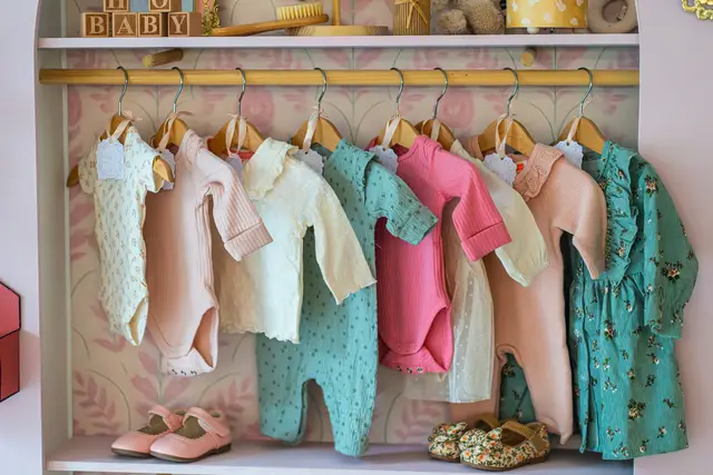 Baby clothes in soft colors hanging on a wooden rail above small shoes