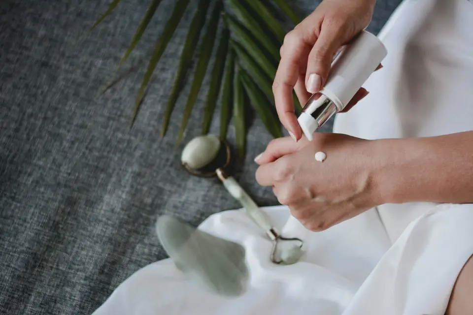 Hands applying lotion beside a jade roller and gua sha stone on a white cloth