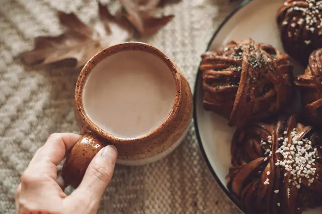 Hand holding a ceramic cup of hot drink beside a plate of pastries and autumn leaves