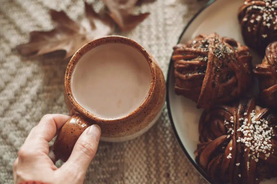 Hand holding a ceramic cup of hot drink beside a plate of pastries and autumn leaves