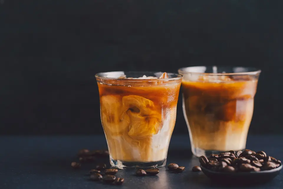 Two glasses of iced coffee with swirls of cream on a dark table scattered with coffee beans