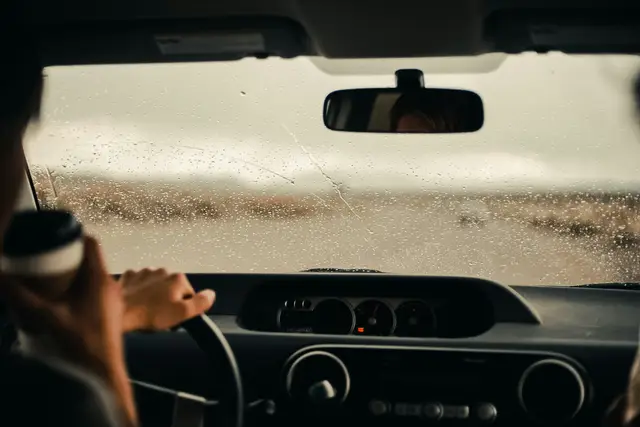 Driver holding a coffee cup with one hand on the steering wheel on a rainy day