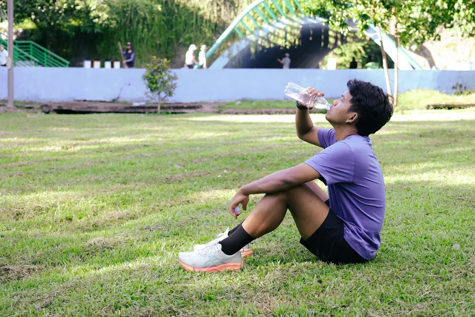 Young man resting on the grass in a park and drinking from a water bottle after a run
