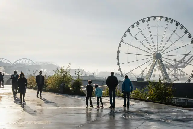 Family and visitors walking along a waterfront promenade past a large ferris wheel