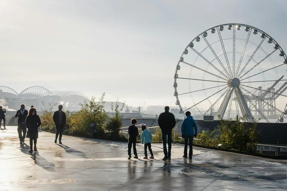 Family and visitors walking along a waterfront promenade past a large ferris wheel