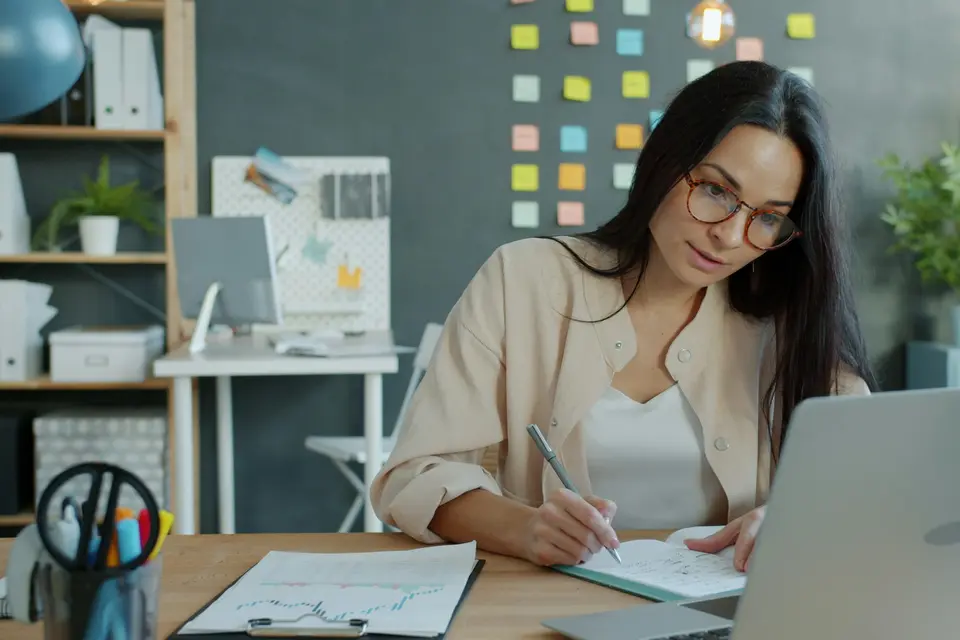 Woman writing notes in a notebook beside an open laptop at a desk