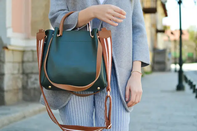 Woman on a city sidewalk carrying a green leather handbag with a tan strap